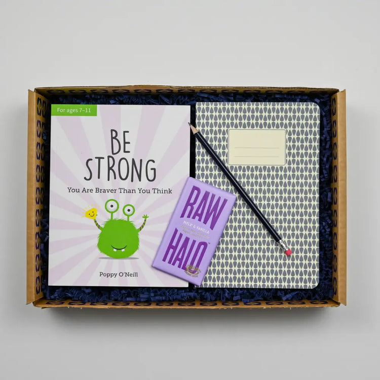 Comforting Care Package for Kids containing a 'BE STRONG' children's book, a purple notebook, and a pencil, all displayed on a bed with a gray blanket.