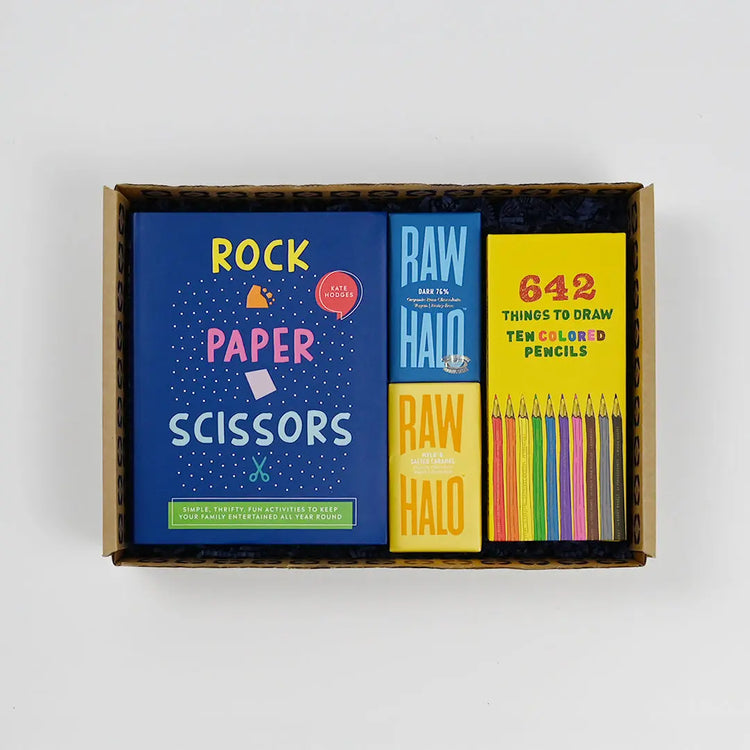 A gift box containing children's art and craft items including a 'ROCK PAPER SCISSORS' game, a 'RAW HALO' drawing challenge, and a set of colored pencils.