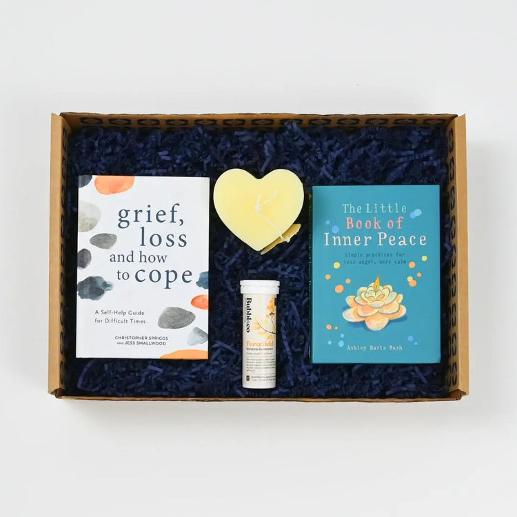 A gift box containing a book titled 'grief, loss and how to cope', forecfield Effervescence, and a decorative heart-shaped candle, all presented on a blue sizzle with gift box.