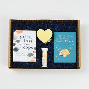 A gift box containing a book titled 'grief, loss and how to cope', forecfield Effervescence, and a decorative heart-shaped candle, all presented on a blue sizzle with gift box.