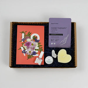 A gift package containing various items including a floral notebook, a box of chocolates, a candle, and skincare products, all arranged in a box with a 'Hand Therapy' packet visible.