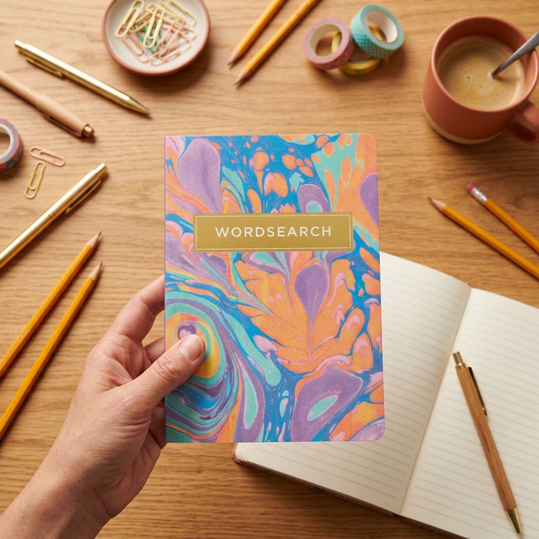 Colorful wordsearch book held by a hand on a desk with stationery items.