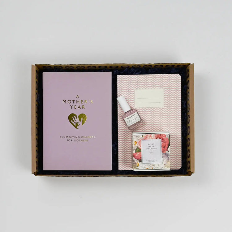 A gift box open to reveal a pink journal, a small bottle of perfume, and a packaged candle, all arranged neatly inside.