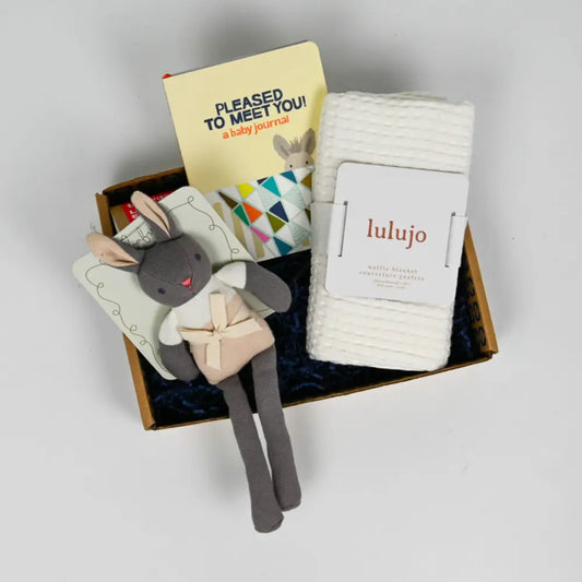 A gift box containing a gray plush toy, a white and gray striped baby blanket, a greeting card, and other baby items.