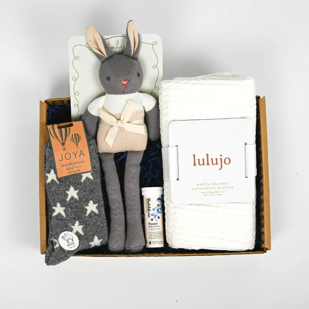 A gift box open to reveal a plush toy, socks, and other items for a newborn baby and mum, presented in a neutral color scheme.