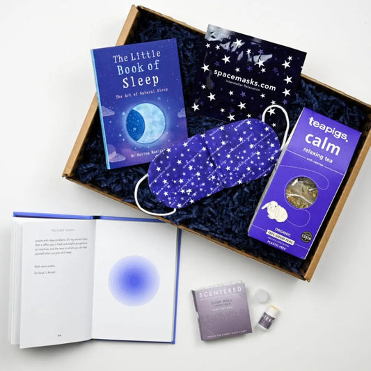 A care package containing a sleep book, a sleeping facemask, tea bags, and a sleep balm, all laid out in a gift box.