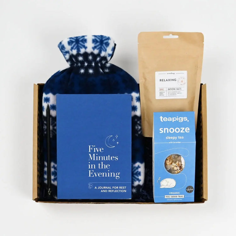 A stress relief gifts package containing a blue hot water bottle cover with white patterns, bath salts with a box of teapigs snooze tea bags, and a blue book titled 'Five Minutes in the Evening'.