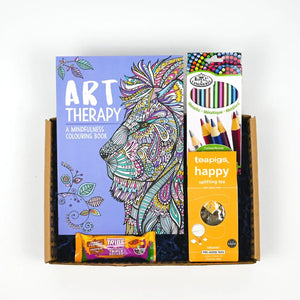 A gift box containing an art therapy colouring book, colouring pencils, a packet of teapigs happy tea and a snack bar.
