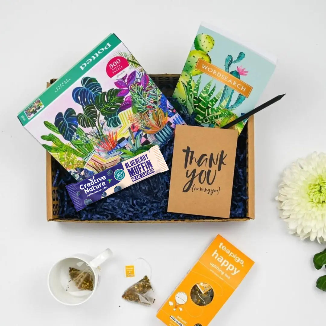 A gift box care package with various items including a colorful potted jigsaw, a wordsearch, teapigs happy tea, a natural snack bar, all arranged on a white background with a flower and a cup of tea.