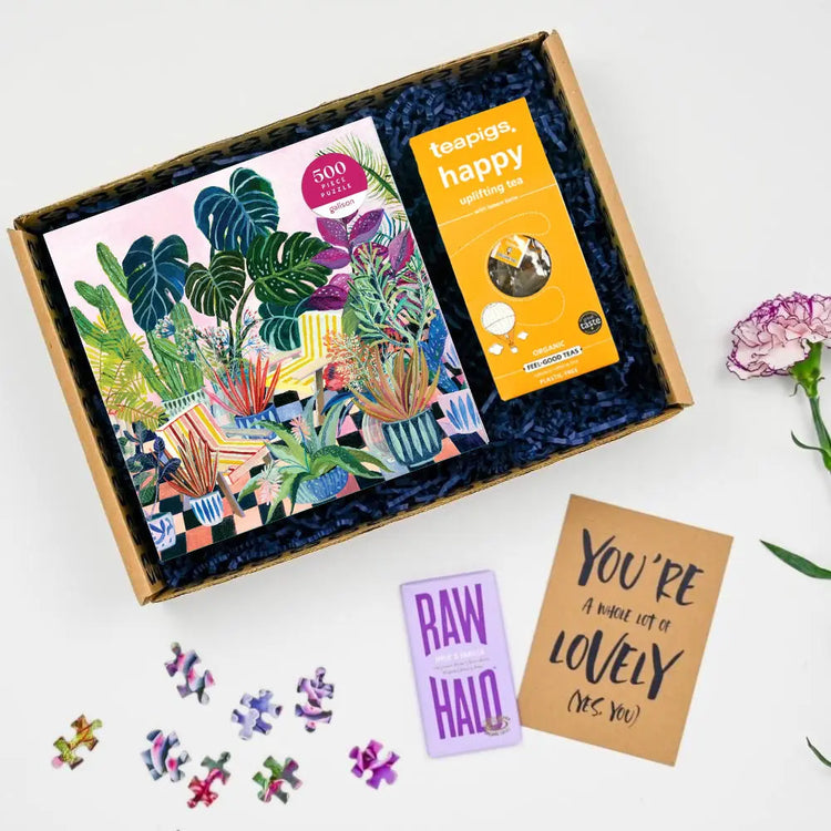 A get well puzzle care package with a colorful jigsaw puzzle box, puzzle pieces, a tea bag, chocolate, and a greeting card displayed on a white background.