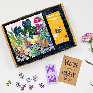 A get well puzzle care package with a colorful jigsaw puzzle box, puzzle pieces, a tea bag, chocolate, and a greeting card displayed on a white background.