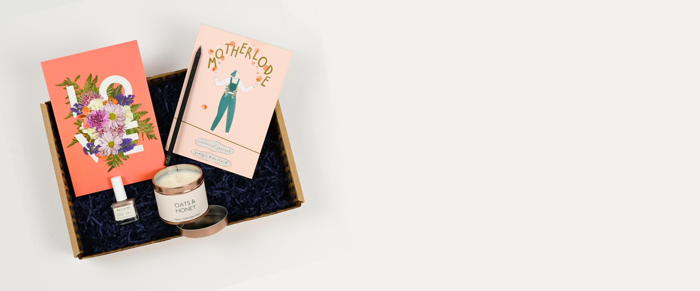 A curated gift box set for her on an off-white background, featuring a coral-pink notebook, a light pink 'MOTHERLODE' book, a small Oats and honey candle tin, and some nailkind nail varnish arranged in a brown kraft gift box with dark blue filler.