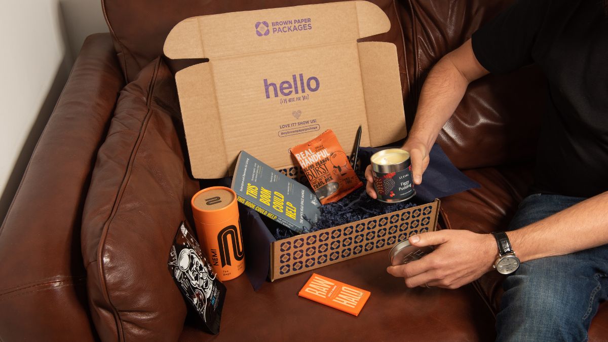 Man unpacking a care package from brown paper packages on a brown leather couch with various gift items including a candle and a power bank.
