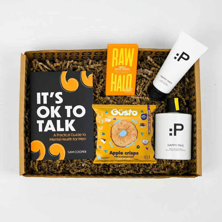 A gift package containing a book titled 'It's Ok to Talk', a packet of apple crisps, and some Happy Paul aromatherapy product, all arranged in a gift box.