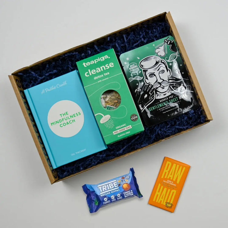 A Men's Mindfulness Care Package gift box containing various items including a book, a packet of tea, and a chocolate bar.