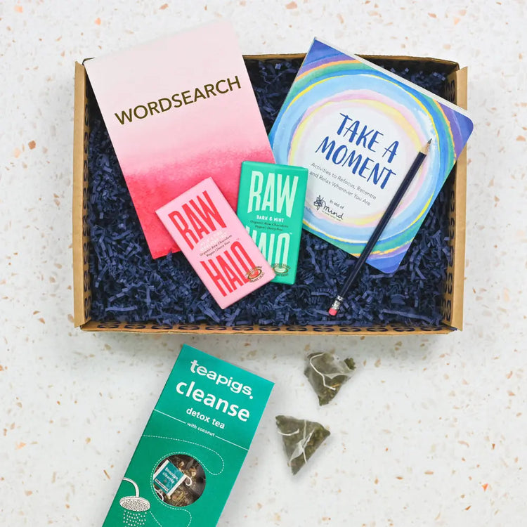 A care package containing various items for relaxation such as a word search book, a wellbeing book, tea and treats, displayed in an open gift box on a kitchen counter.