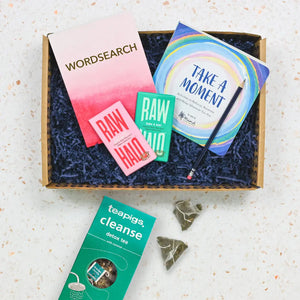 A care package containing various items for relaxation such as a word search book, a wellbeing book, tea and treats, displayed in an open gift box on a kitchen counter.
